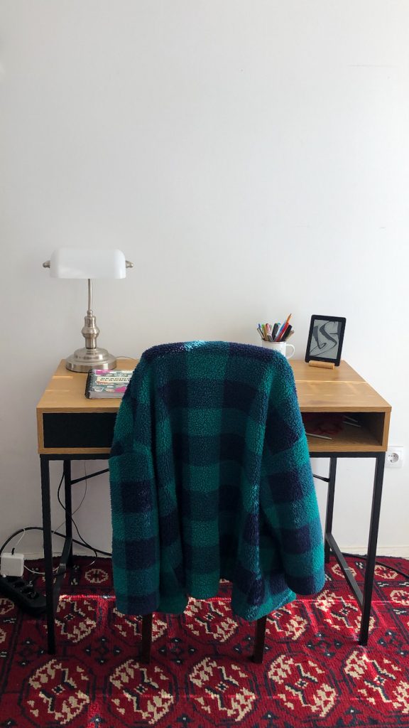 A picture of wooden desk on the red bokhara oriental rug. Green checkered cardigan is draped over the chair. On the desk there is white banker's lamp, Kindle, journal and cup with pens. 